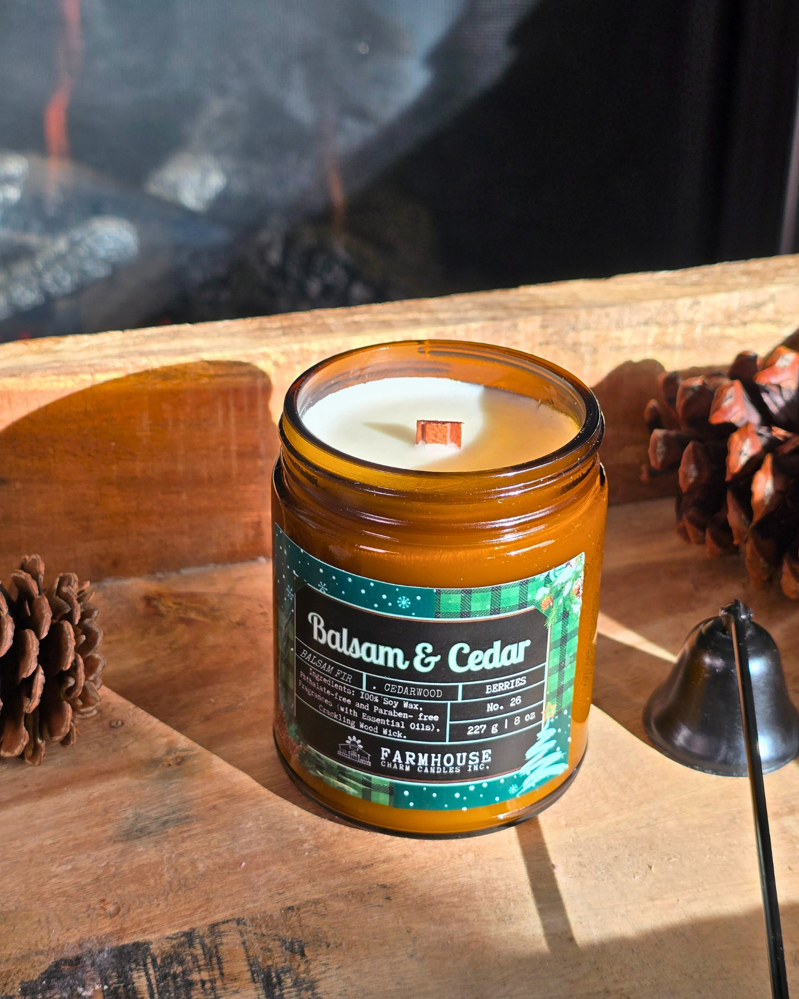 Candle in a glass jar labeled 'Balsam & Cedar' by Farmhouse Charm Candles on a wooden surface with pinecones.