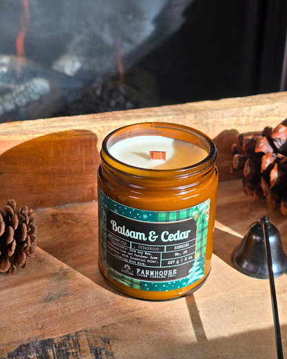 Candle in a glass jar labeled 'Balsam & Cedar' by Farmhouse Charm Candles on a wooden surface with pinecones.