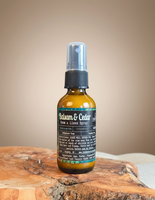 Amber bottle of Balsam & Cedar Room & Linen Spray by Farmhouse Charm Candles, styled on a cozy winter backdrop with pine branches and berries, evoking a fresh woodsy and cabin-like scent.
