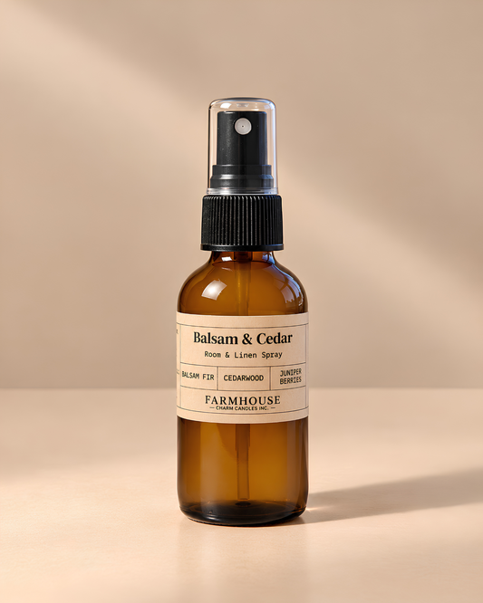 An amber glass room and linen spray bottle with a black spray top and a cream label reading 'Balsam & Cedar' and 'FARMHOUSE Charm Candles', photographed on a warm beige background with soft shadows.