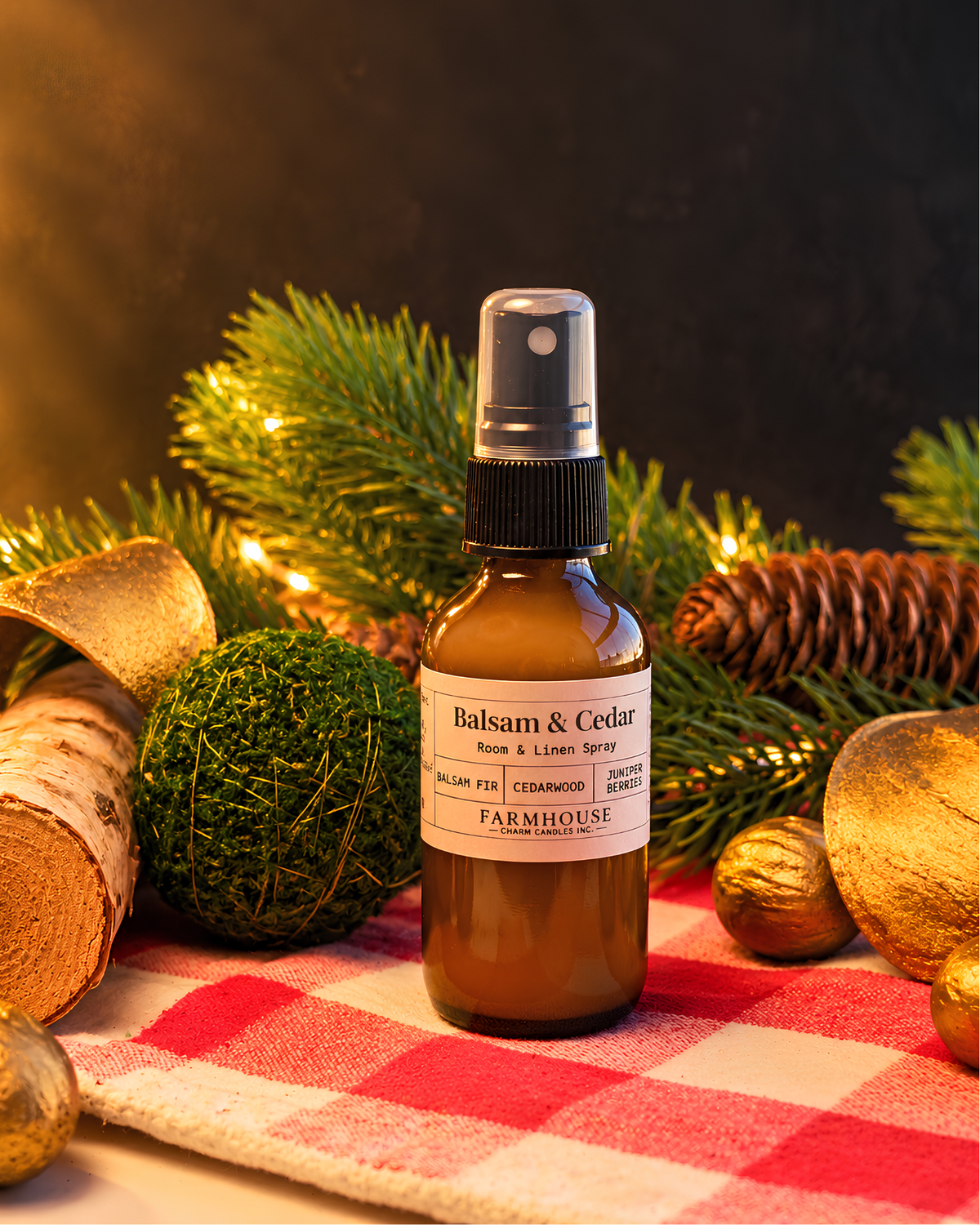 A brown glass spray bottle labeled 'Balsam & Cedar' by Farmhouse Charm Candles, styled on a red-and-white checkered cloth with pine branches, a moss ball, and gold ornaments against a dark background.