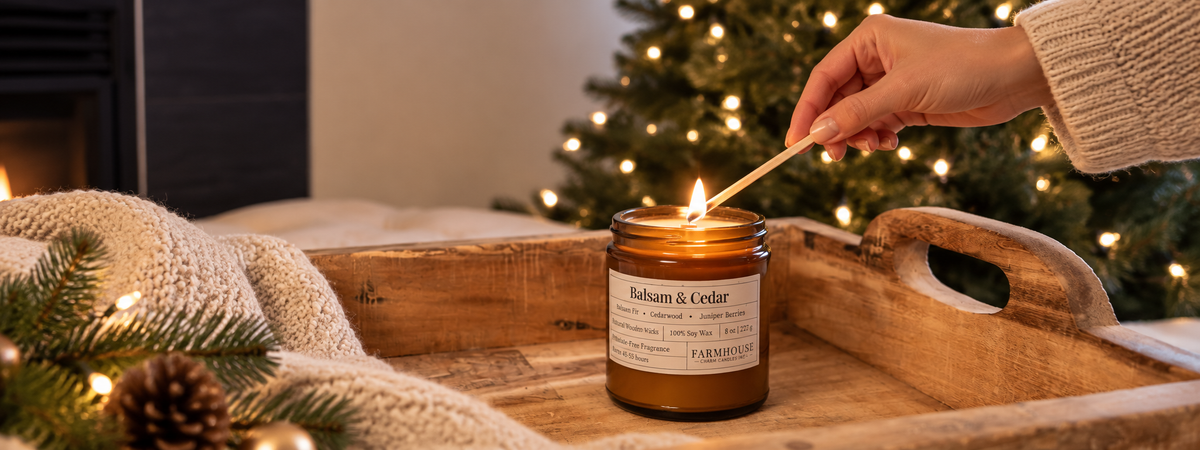 A lit amber glass candle on a wooden tray, with a hand holding a match above it. A cozy holiday scene with a blurred Christmas tree, blanket, and fireplace in the background. Home for Christmas by Farmhouse Charm Candles