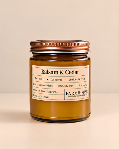 A Farmhouse Charm Candles Balsam & Cedar scented soy wax candle in a clear glass jar with a copper-toned metal lid, set against a solid beige background.