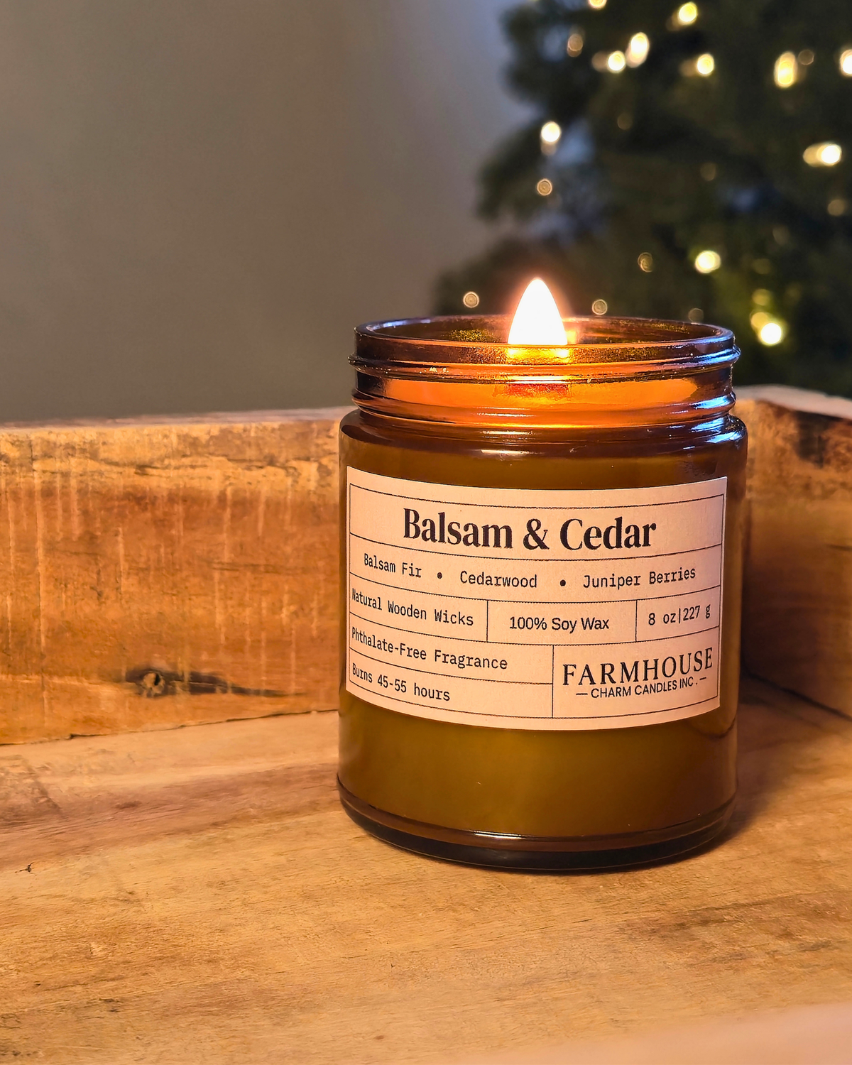 A lit amber glass jar candle labeled 'Balsam & Cedar' by Farmhouse Charm Candles ., placed on a rustic wooden surface with a blurred Christmas tree in the background.