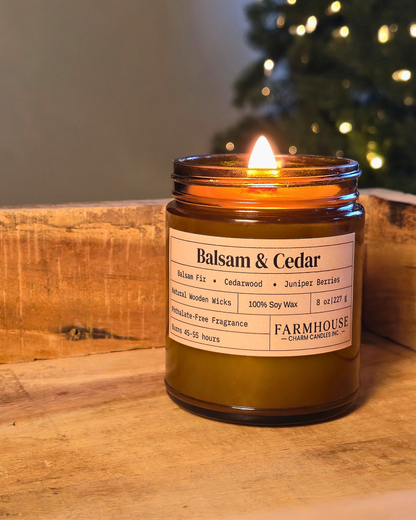 A lit amber glass jar candle labeled 'Balsam & Cedar' by Farmhouse Charm Candles ., placed on a rustic wooden surface with a blurred Christmas tree in the background.