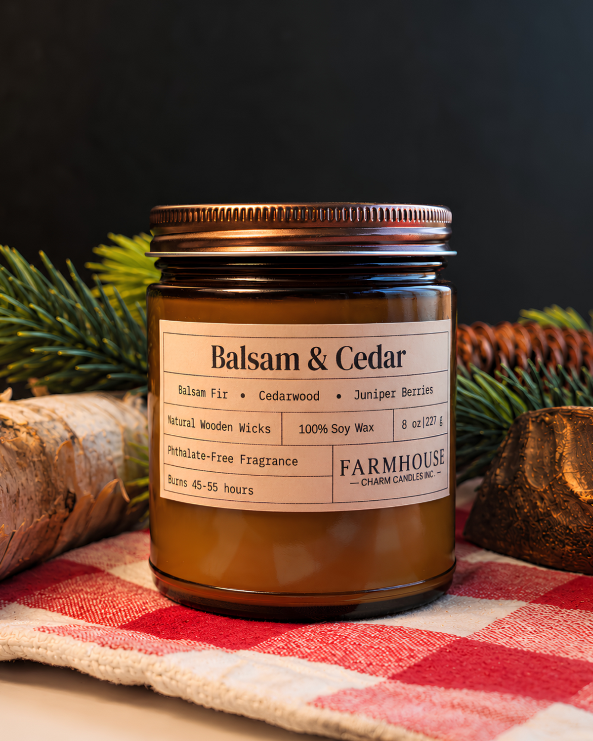 A  Farmhouse Charm Candle jar candle with Balsam & Cedar scent, styled on a red and white checkered cloth with pine branches, birch bark, and wood pieces against a dark background.