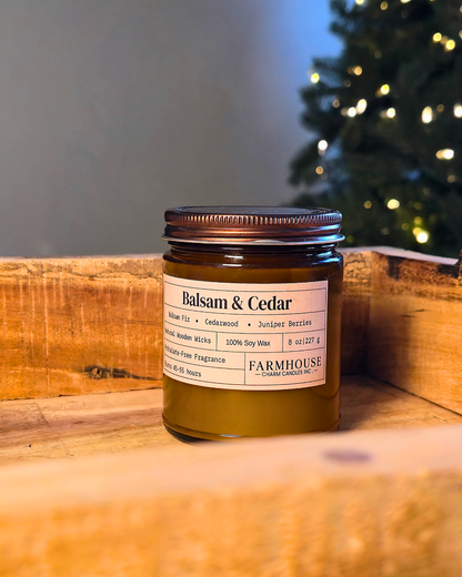 A jar candle labeled 'Balsam & Cedar' by  Farmhouse Charm Candles placed on a wooden surface with a blurred Christmas tree and warm indoor background.