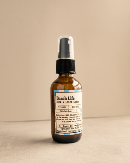 Brown spray bottle labeled 'Beach Life' by Farmhouse Charm Candles on a gray surface with a warm background
