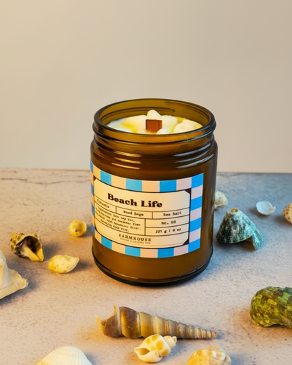 Candle in a jar labeled 'Beach Life' by Farmhouse Charm Candle with seashells and stones on a sandy surface.