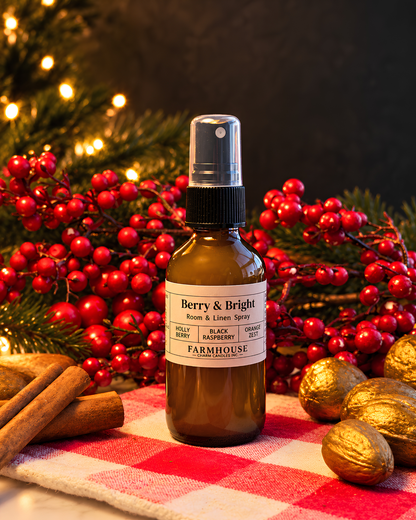 A brown glass room and linen spray bottle with a black spray top and a white label reading 'Berry & Bright' by 'FARMHOUSE Charm candles styled on a red-and-white checkered cloth with cinnamon sticks, gold pinecones, and red berries against a dark background with blurred holiday lights.