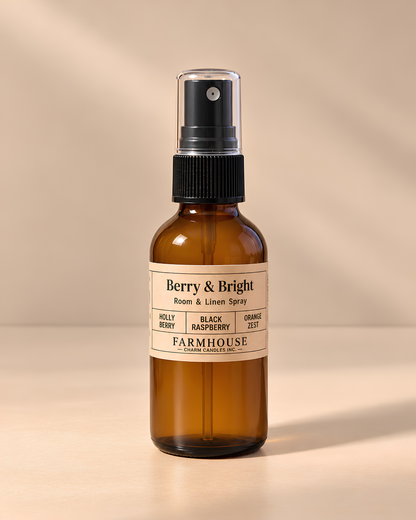 An amber glass room and linen spray bottle with a black spray top and a cream label reading 'Berry & Bright' by FARMHOUSE Charm Candles photographed on a warm beige background with soft shadows.