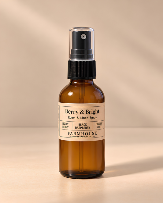 An amber glass room and linen spray bottle with a black spray top and a cream label reading 'Berry & Bright' by FARMHOUSE Charm Candles photographed on a warm beige background with soft shadows.
