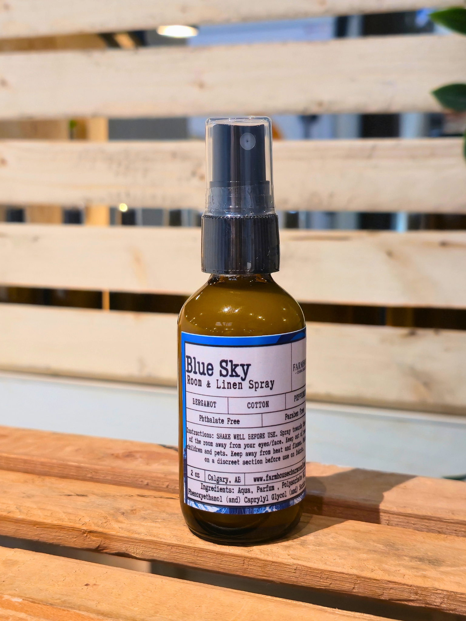 Bottle of Blue Sky Room & Linen Spray by Farmhouse Charm Candles on a wooden surface with a blurred background