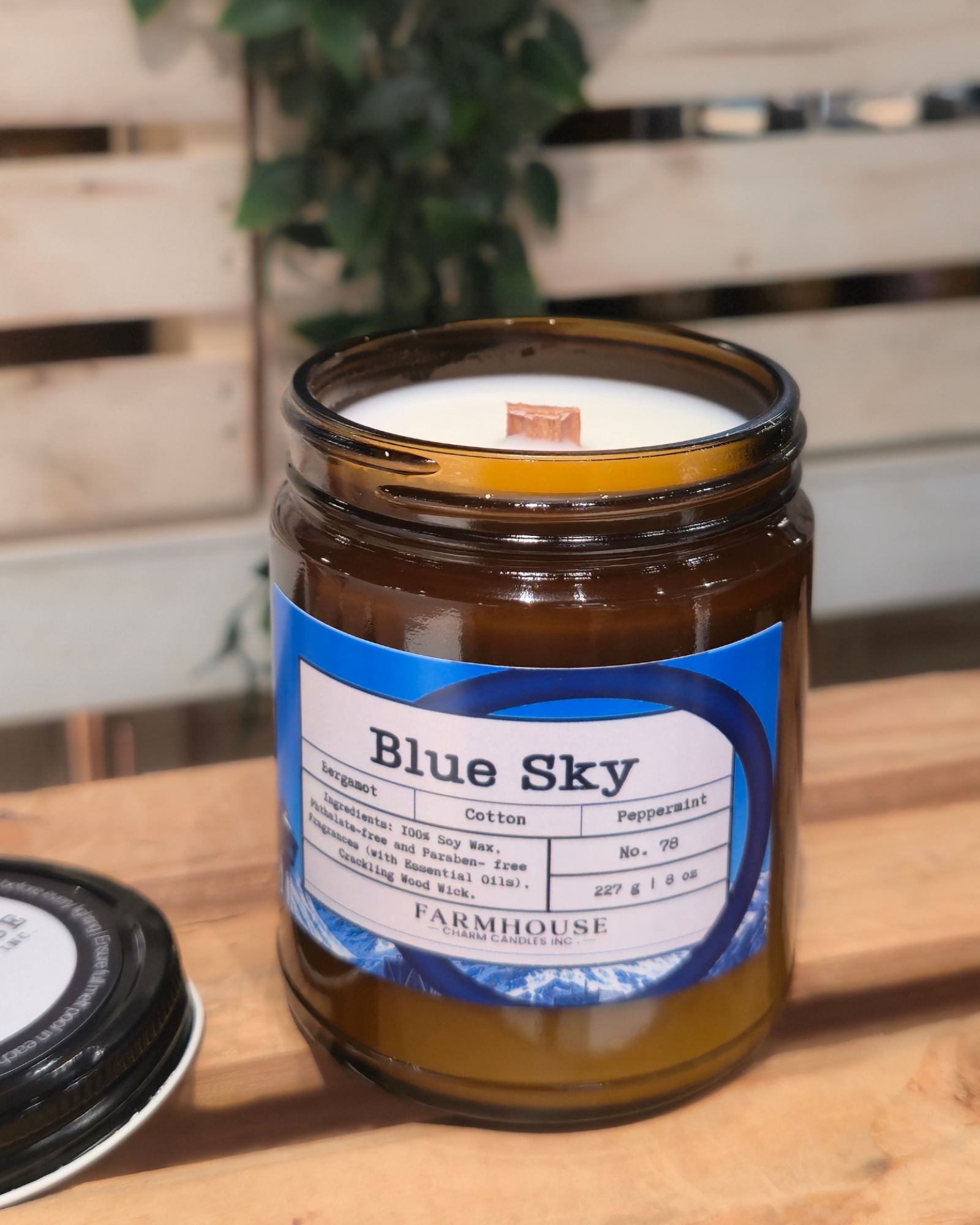Candle in a glass jar labeled 'Blue Sky' by Farmhouse Charm Candles on a wooden surface with a plant in the background.