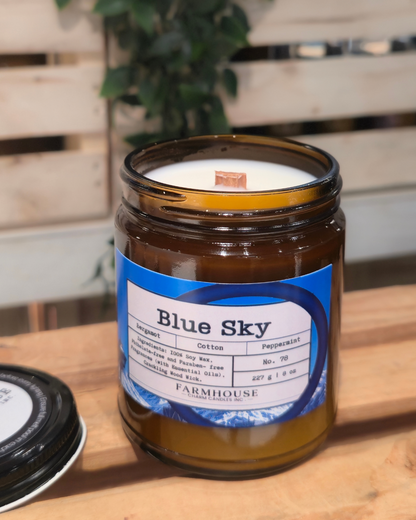 Candle in a glass jar labeled 'Blue Sky' by Farmhouse Charm Candles on a wooden surface with a plant in the background.