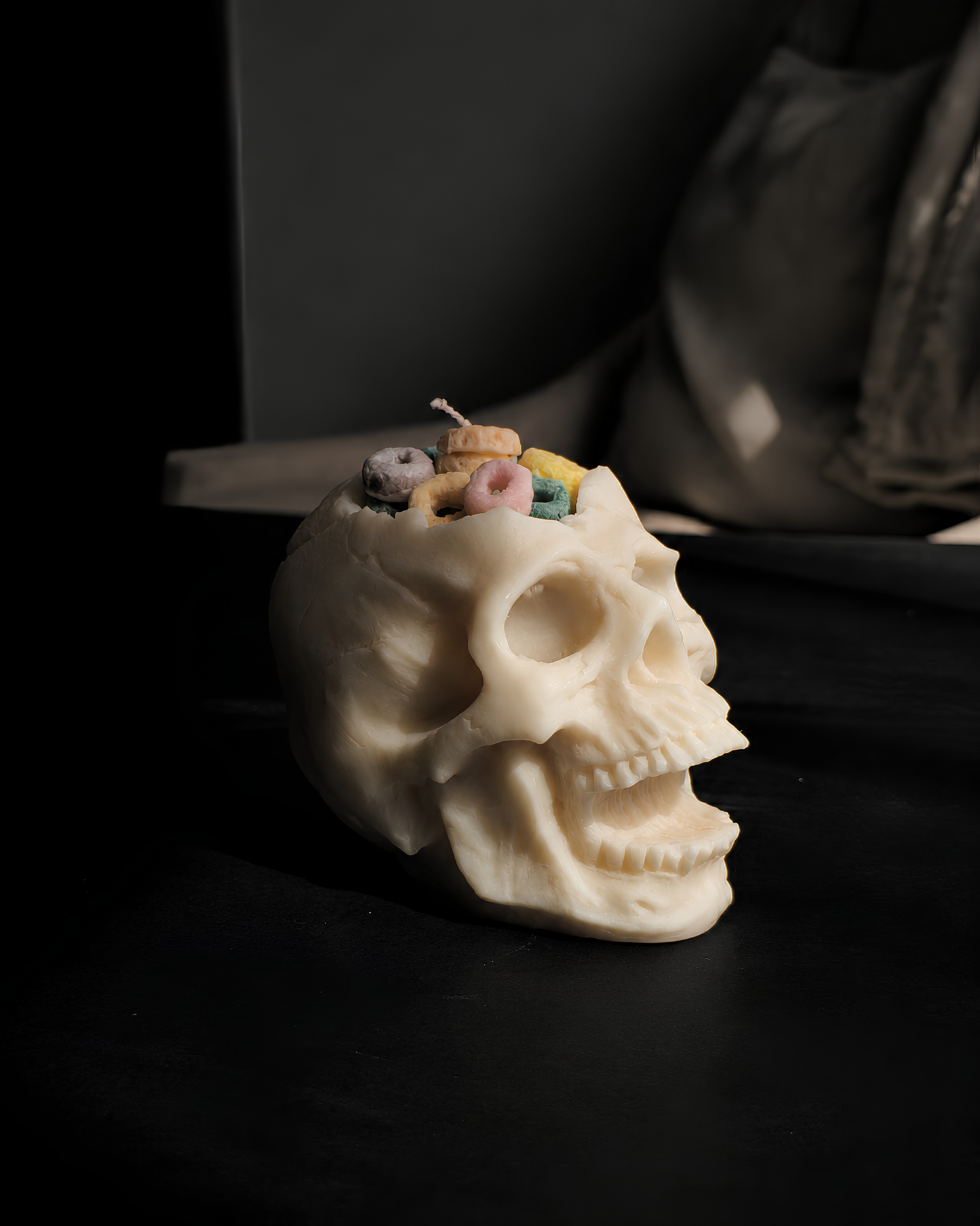A cream-colored skull-shaped candle with colorful candy-like details inside the cranium, set on a dark surface with a moody, dimly lit background. Cereal Killer Pillar Candle by Farmhouse Charm Candles