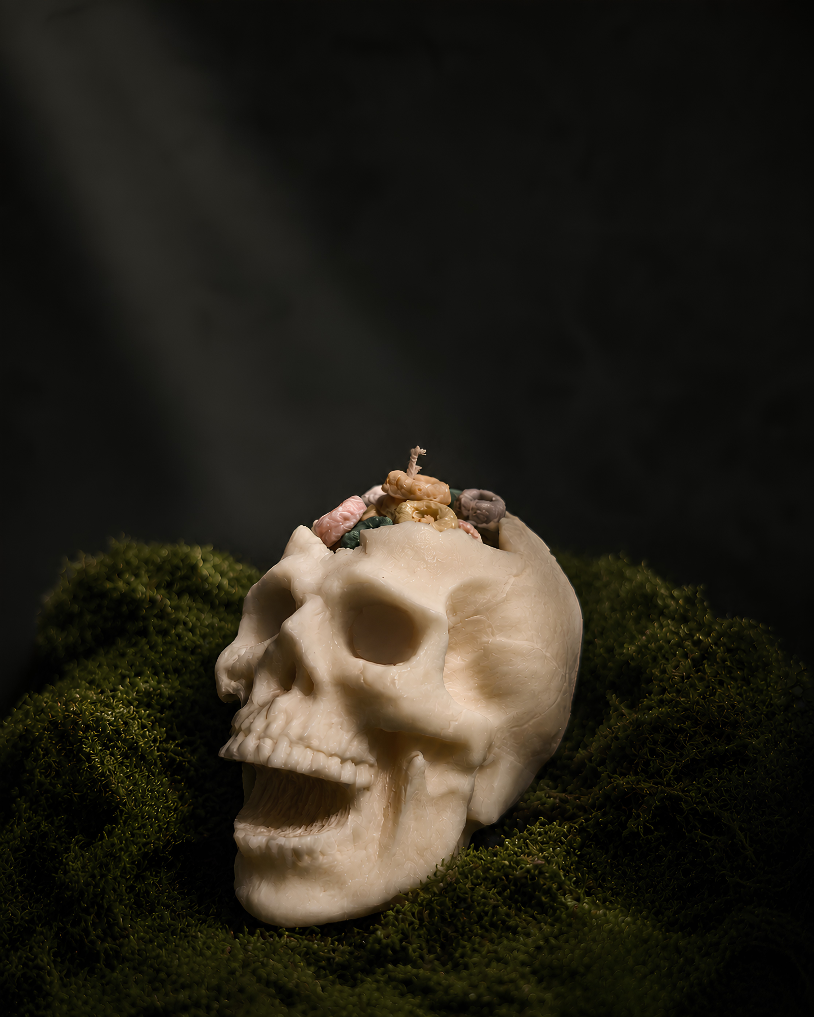 A cream-colored skull sculpture resting on a bed of dark green moss against a dark, moody background with dramatic light rays. Cereal Killer Pillar Candle by Farmhouse Charm Candles