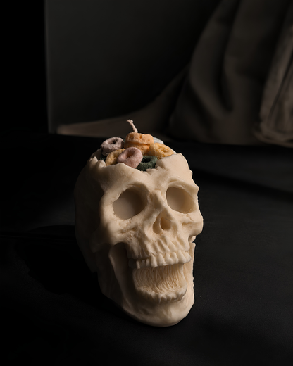 An off-white skull-shaped candle with a wick on top and small pastel decorative accents, photographed on a dark black surface with a soft shadow. Cereal Killer Pillar Candle by Farmhouse Charm Candles