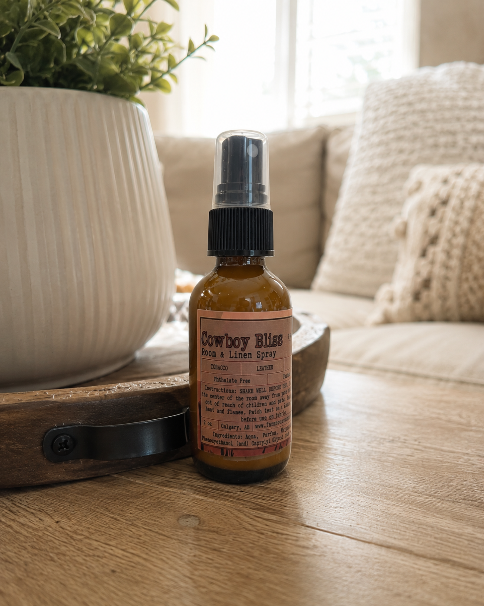 Cowboy Bliss Room and Linen Spray amber bottle on rustic wood tray beside a white ceramic planter, cozy living room setting with neutral sofa in background — Farmhouse Charm Candles