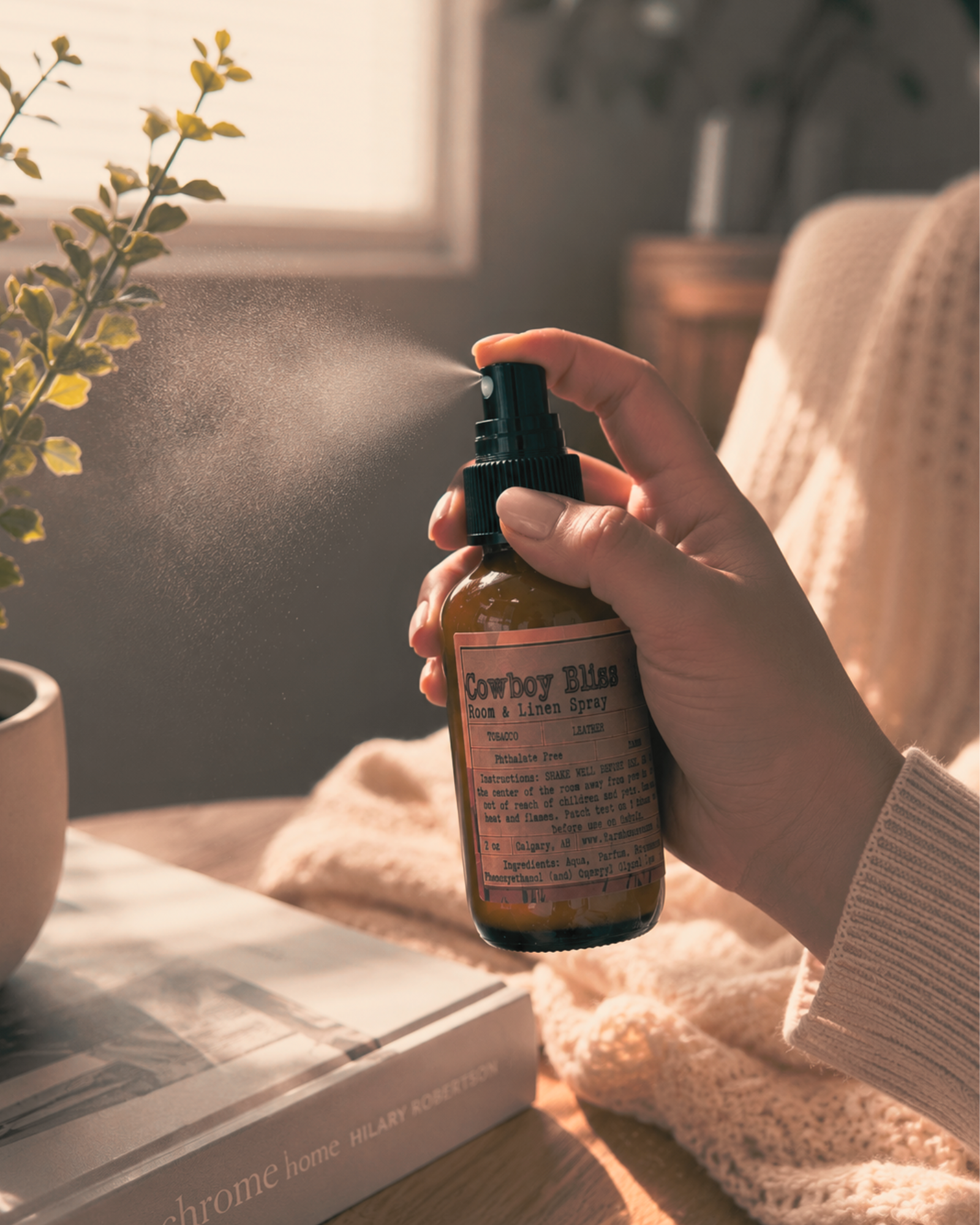 Hand spraying Cowboy Bliss Room and Linen Spray with visible mist, warm sunlit home setting with knit blanket, books and green plant — Farmhouse Charm Candles