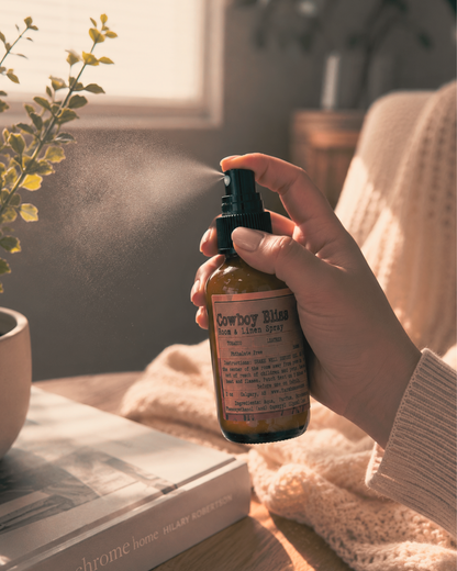 Hand spraying Cowboy Bliss Room and Linen Spray with visible mist, warm sunlit home setting with knit blanket, books and green plant — Farmhouse Charm Candles