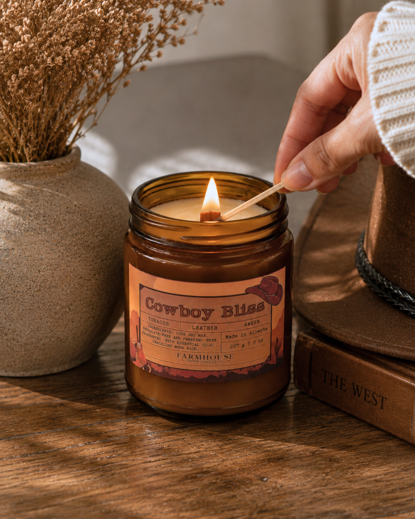 A hand lights the wooden wick of the Cowboy Bliss soy candle in an amber glass jar, set on a rustic wooden table. The warm glow is complemented by a ceramic vase with dried florals, a vintage cowboy hat, and a weathered book in the background, creating a cozy Western farmhouse atmosphere. Farmhouse Charm Candles