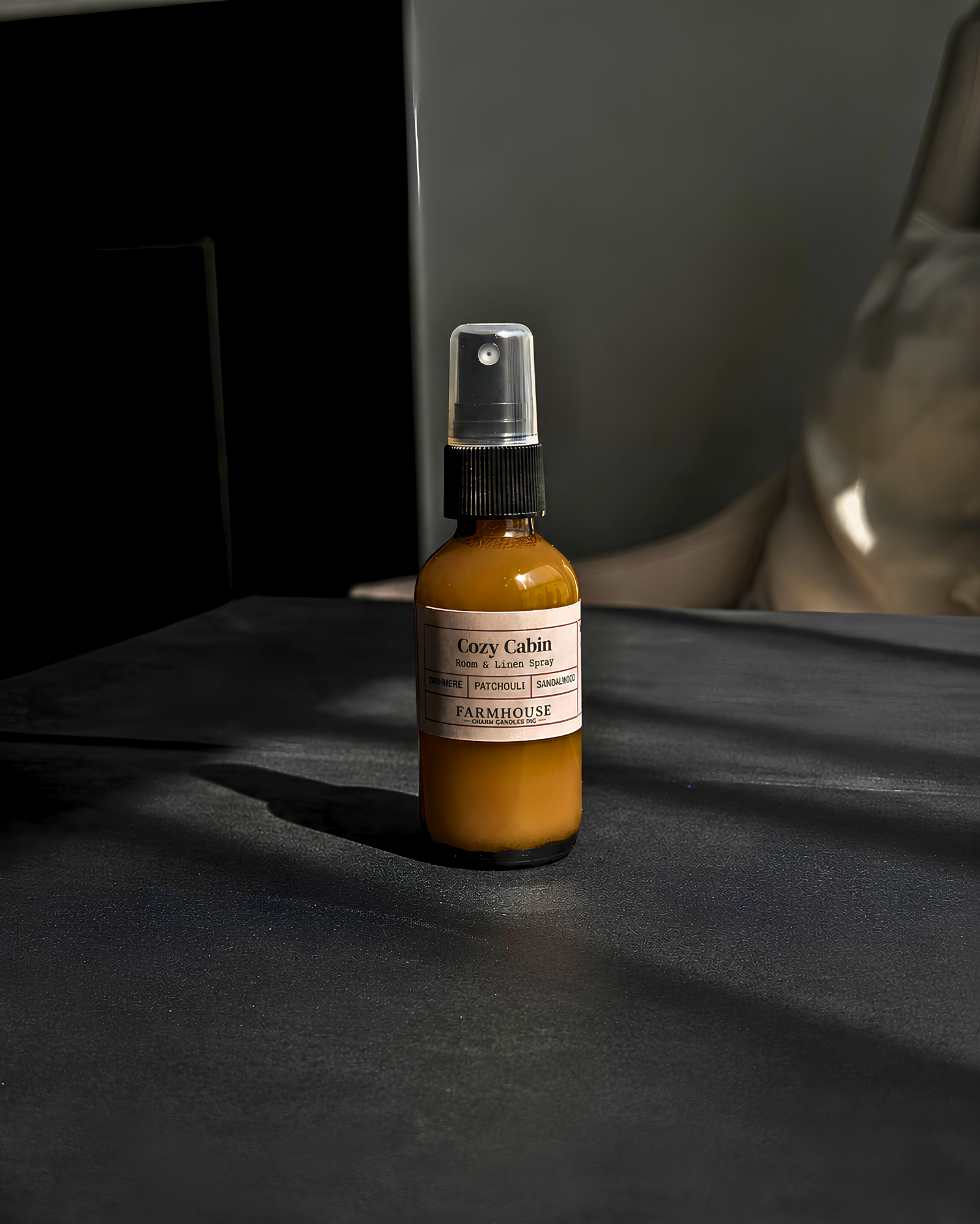 A small amber glass spray bottle with a black spray top and a cream label reading 'Cozy Cabin' by  Farmhouse Charm Candles, standing on a dark tabletop in moody lighting.