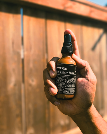 Hand holding a bottle of 'Cozy Cabin' room & linen spray by Farmhouse Charm Candles against a wooden fence background