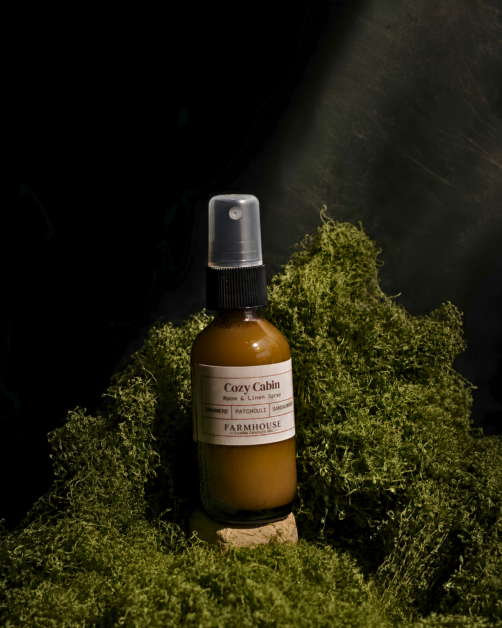 A bottle of Cozy Cabin room & linen spray by  Farmhouse Charm Candles, with a white label and black spray cap, set on a cork against a dark background with green moss.