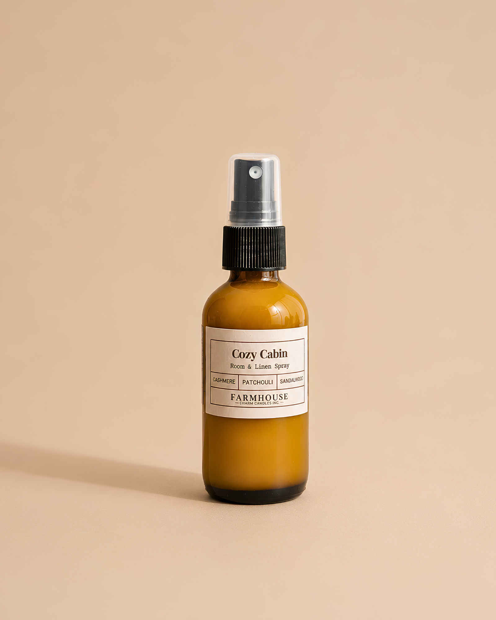 An amber glass spray bottle with a black pump and a cream label reading 'Cozy Cabin' by Farmhouse Charm Candles, centered on a warm beige background.