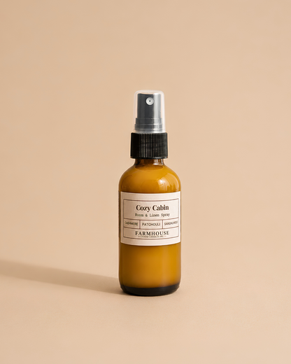 An amber glass spray bottle with a black pump and a cream label reading 'Cozy Cabin' by Farmhouse Charm Candles, centered on a warm beige background.