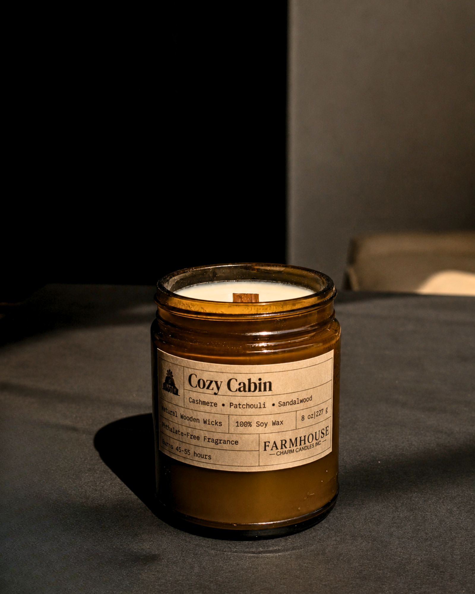 A Farmhouse Charm Candles 'Cozy Cabin' scented soy wax candle in an amber glass jar, set on a dark gray surface with a dark gray background.