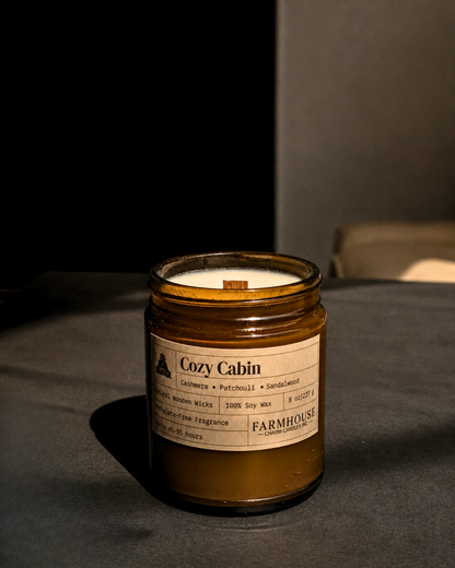 A Farmhouse Charm Candles 'Cozy Cabin' scented soy wax candle in an amber glass jar, set on a dark gray surface with a dark gray background.