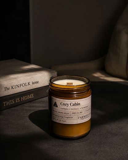 A candle in an amber glass jar with a white label reading 'Cozy Cabin' by  Farmhouse Charm Candles styled on a dark surface beside stacked books in a moody indoor setting.