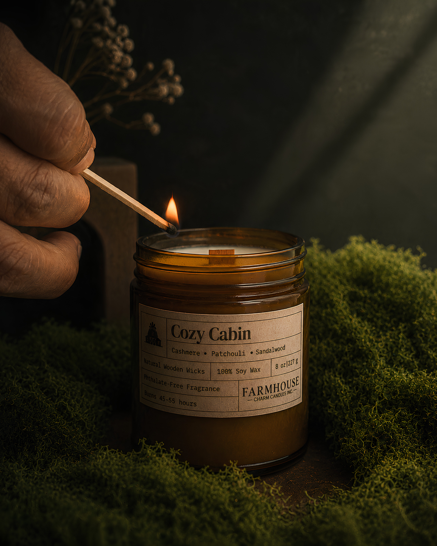A hand holds a lit match to a amber glass jar candle labeled 'Cozy Cabin' by Farmhouse Charm Candles, set on moss with a dark moody background.