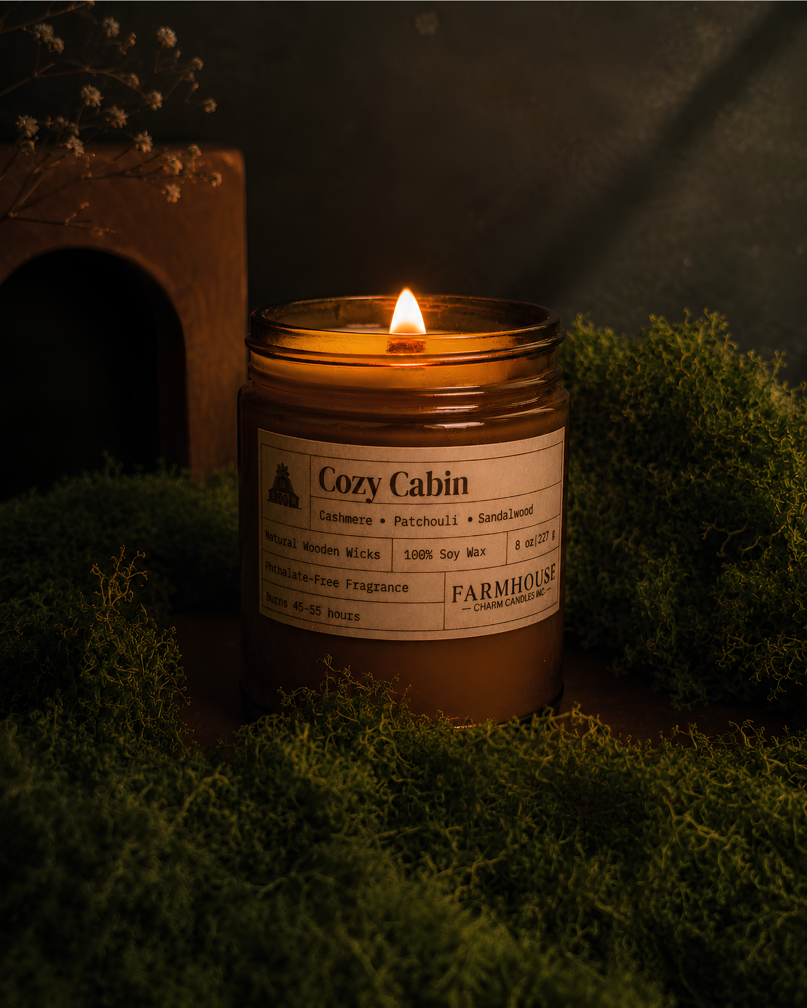 A lit amber glass jar candle labeled 'Cozy Cabin' by  Farmhouse Charm Candles, placed on green moss with a dark rustic background and a small wooden arch in the distance.