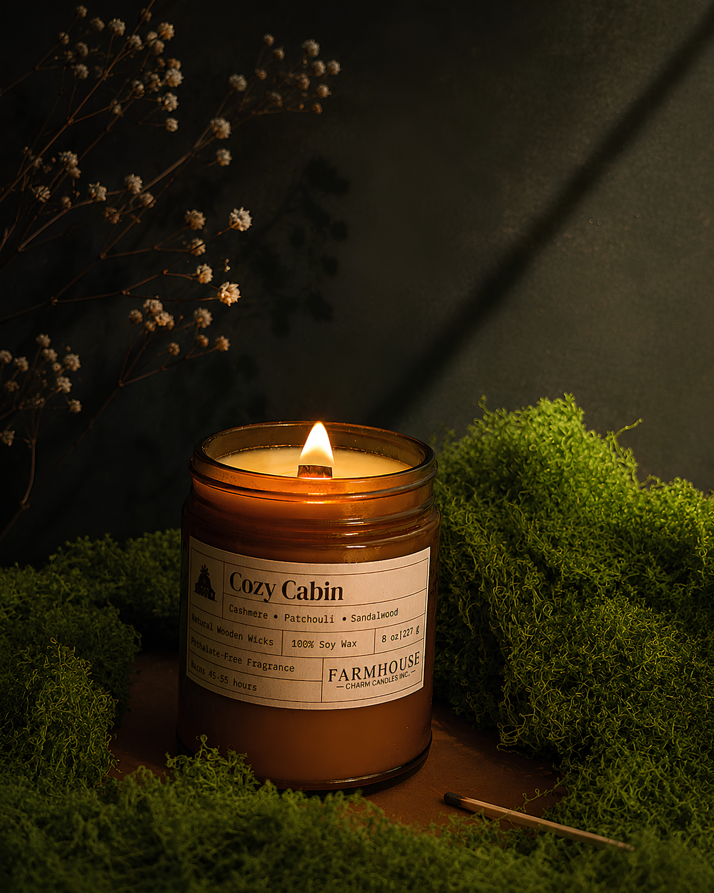 A lit amber glass jar candle labeled 'Cozy Cabin' by Farmhouse Charm Candles styled on moss with dried flowers against a dark background.
