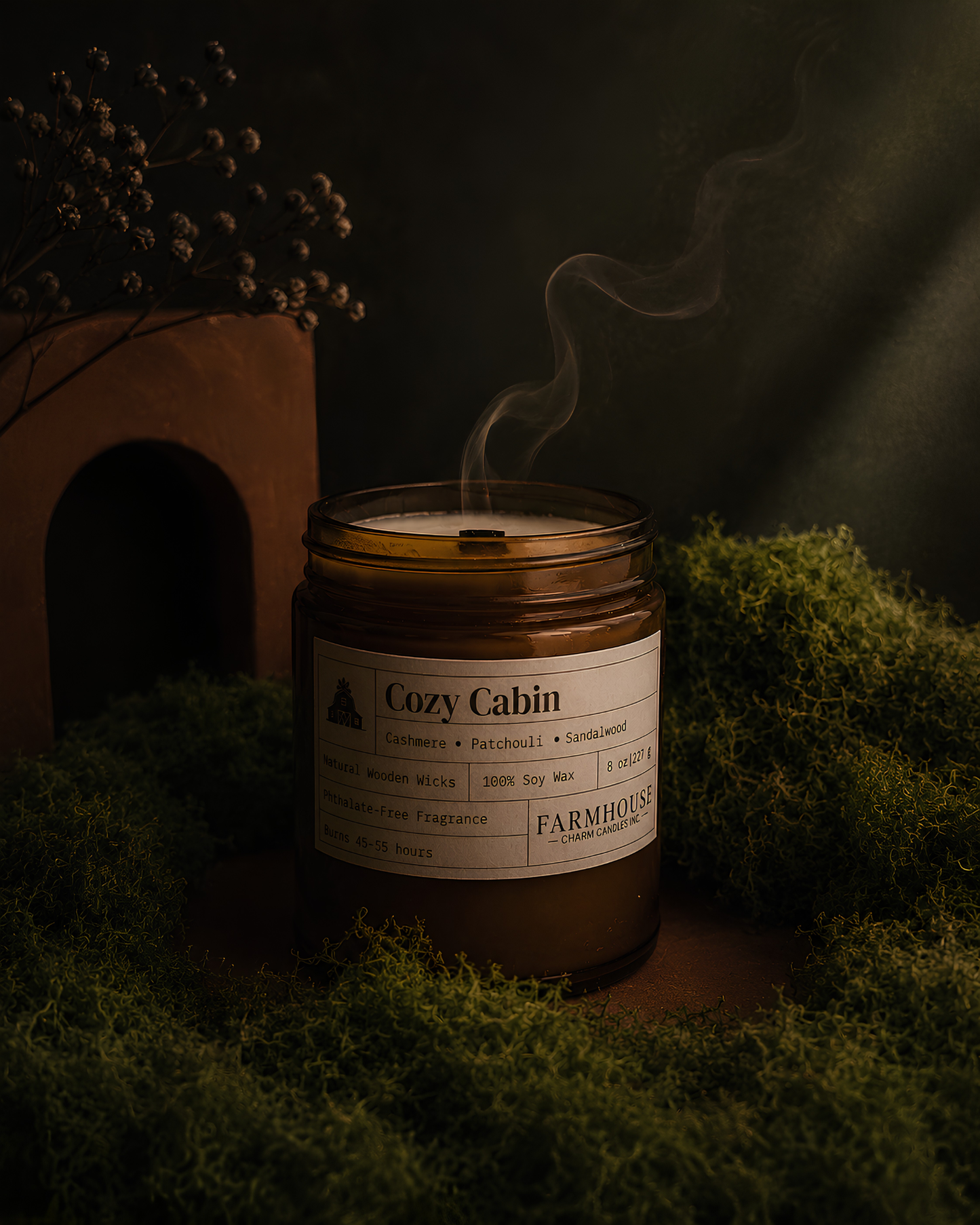 A rustic amber glass jar candle labeled 'Cozy Cabin' sits on moss in a moody, dark setting with a small archway and dried flowers nearby. Steam rises from the candle, and the label reads ' Farmhouse Charm Candles
