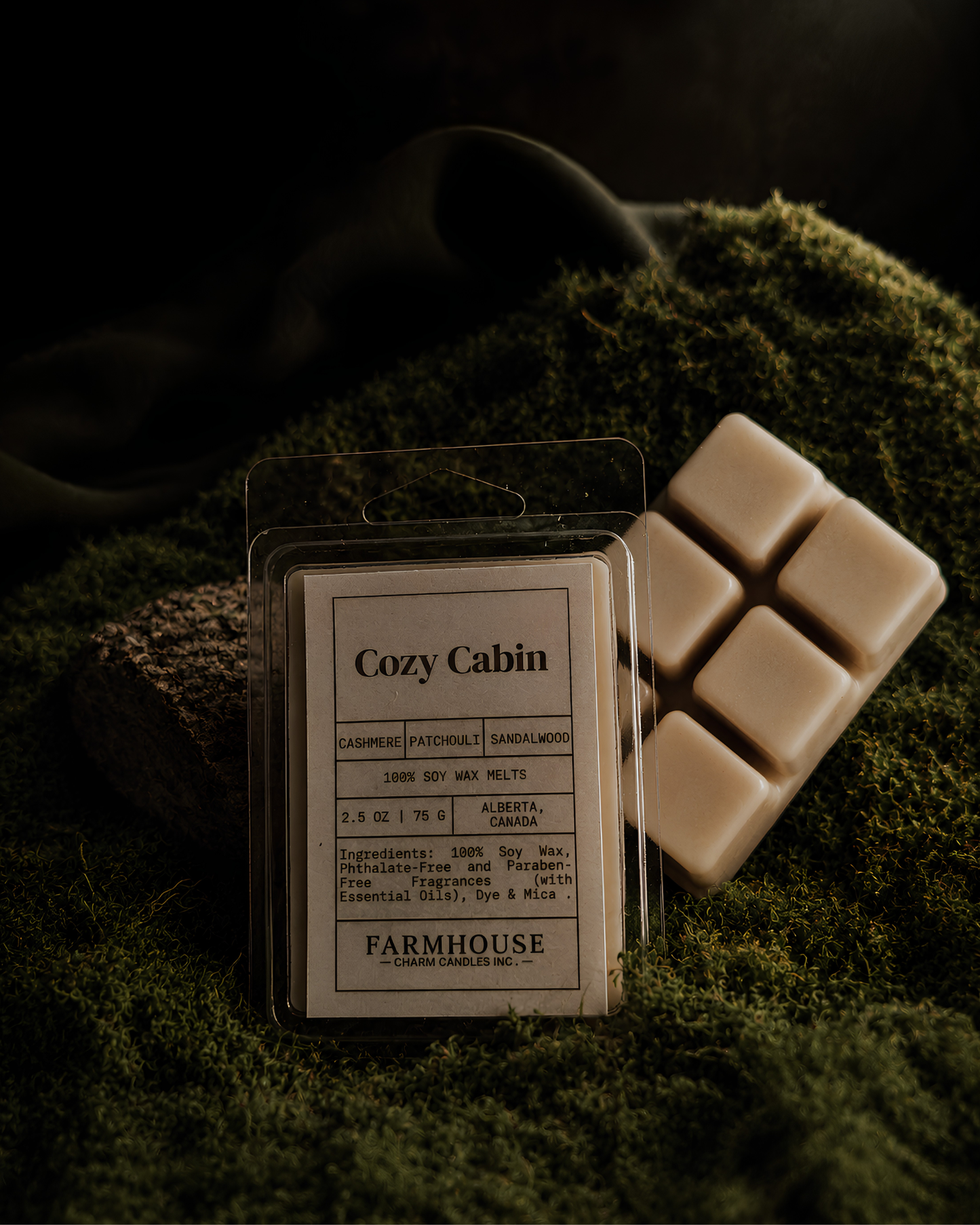 A wax melt package labeled 'Cozy Cabin' by Farmhouse Charm Candles with several cream-colored wax melts beside it, styled on dark green moss against a dark background.