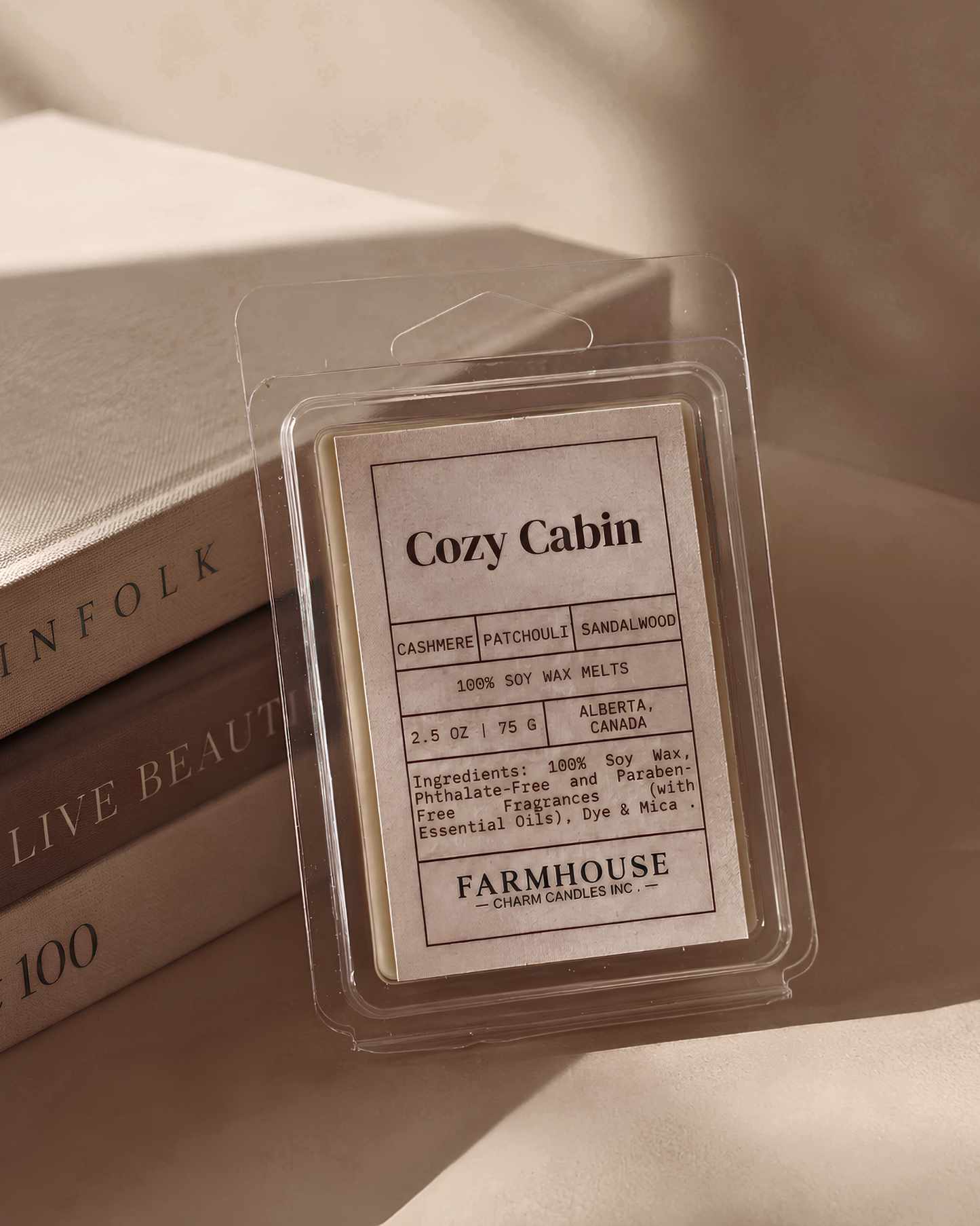 A clear plastic clamshell package containing a Cozy Cabin fragrance wax melt by Farmhouse Charm Candles, styled on top of stacked books with warm, soft lighting.