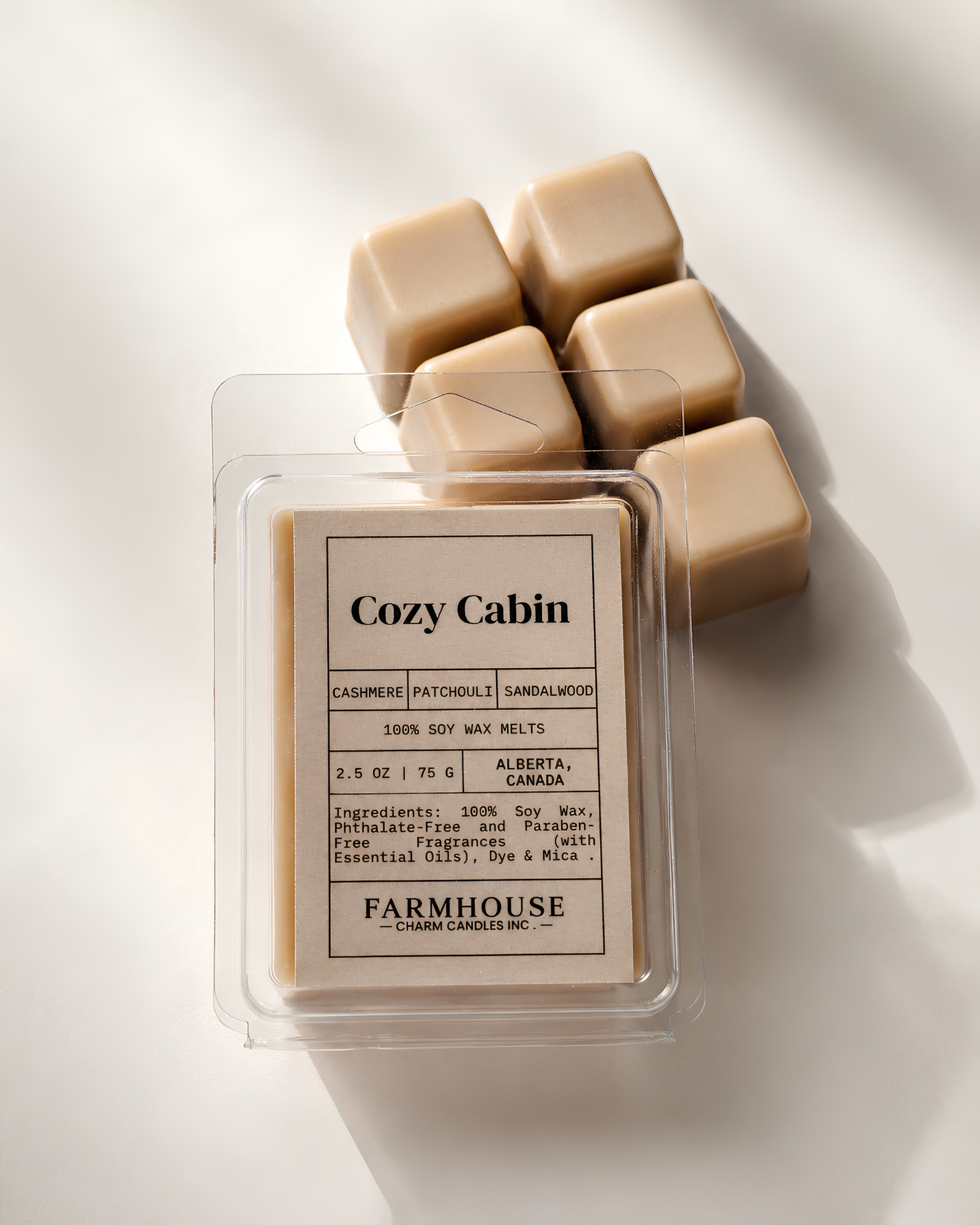 A package of Cozy Cabin soy wax melts  by Farmhouse Charm Candles in a clear clamshell container, with several beige wax cubes arranged behind it on a white background with soft shadows.