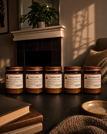Five Farmhouse brand scented candles in amber glass jars with copper lids are lined up on a dark table in a cozy, warmly lit room with a fireplace, books, and a knit blanket. Fall Candles Collection by Farmhouse Charm Candles