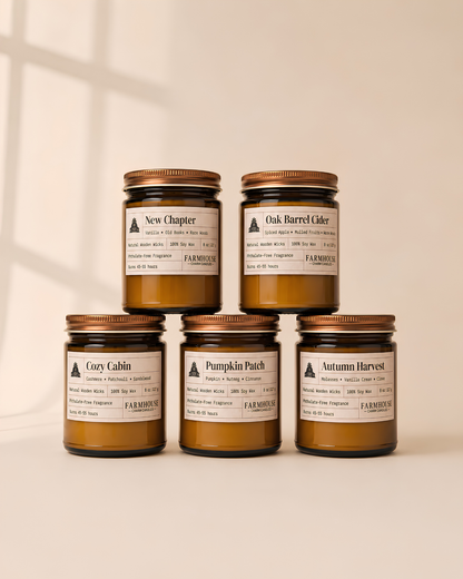 Five amber glass jar candles with gold lids and cream labels are arranged in a pyramid on a light beige surface. The labels show different scent names including New Chapter, Oak Barrel Cider, Cozy Cabin, Pumpkin Patch, and Autumn Harvest. Fall Candles Collection by Farmhouse Charm Candles