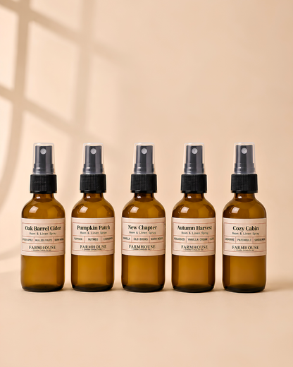 Five amber glass spray bottles with black sprayers and cream labels lined up on a warm beige background. The Fall Collection room & Linen spray by Farmhouse Charm Candles include scents: Oak Barrel Cider, Pumpkin Patch, New Chapter, Autumn Harvest, and Coy Cabin