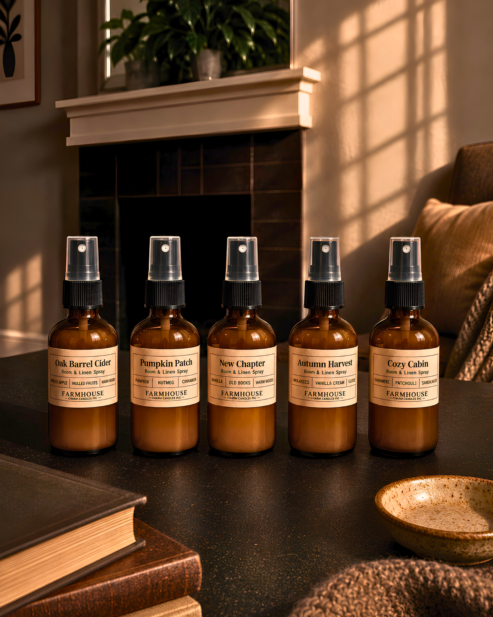 Five amber spray bottles with cream labels lined up on a dark tabletop in a warm, sunlit room. The labels read Oak Barrel Cider, Pumpkin Patch, New Chapter, Autumn Harvest, and Cory Cabin. Fall Collection room & Linen spray by Farmhouse Charm Candles