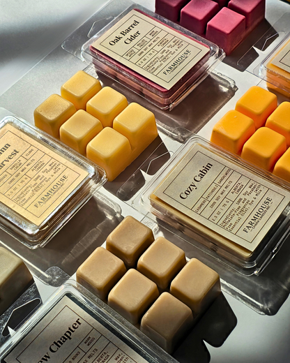 Multiple wax melts in clear plastic clamshell packages arranged on a reflective surface. The wax melts come in warm tones including yellow, tan, cream, and pink, with visible labels reading brands such as FARMHOUSE Charm Candles and scent names like Cozy Cabin, Oak Barrel Cider, and New Chapter.