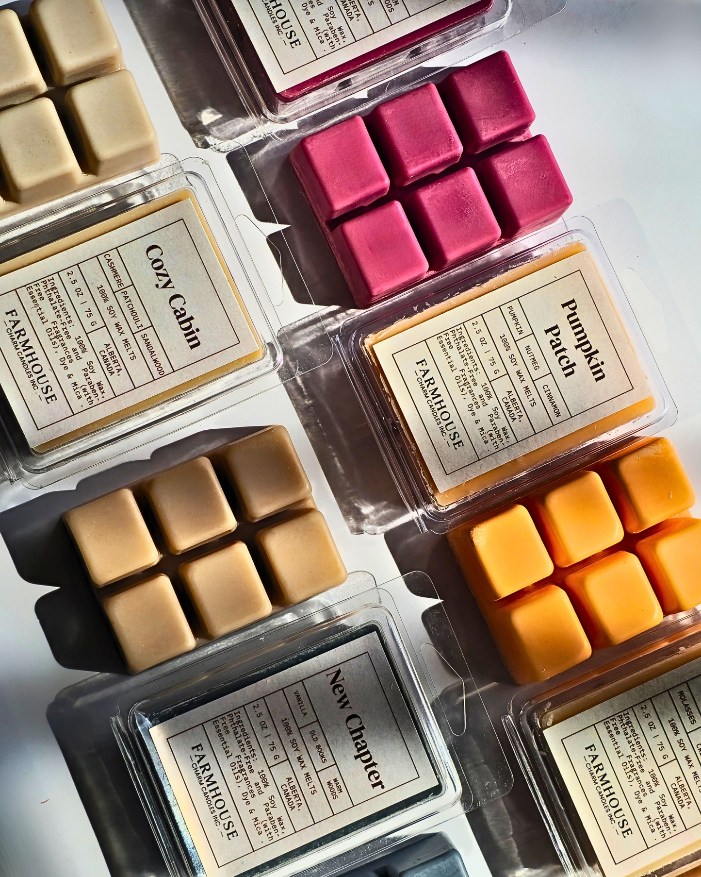 An overhead flat-lay of multiple wax melts in clear clamshell containers with cream labels. Visible scents include Cozy Cabin, Pumpkin Patch, and New Chapter, showing warm beige, pink, tan, and orange wax cubes. Fall Wax Melts Bundle by FARMHOUSE Charm Candles