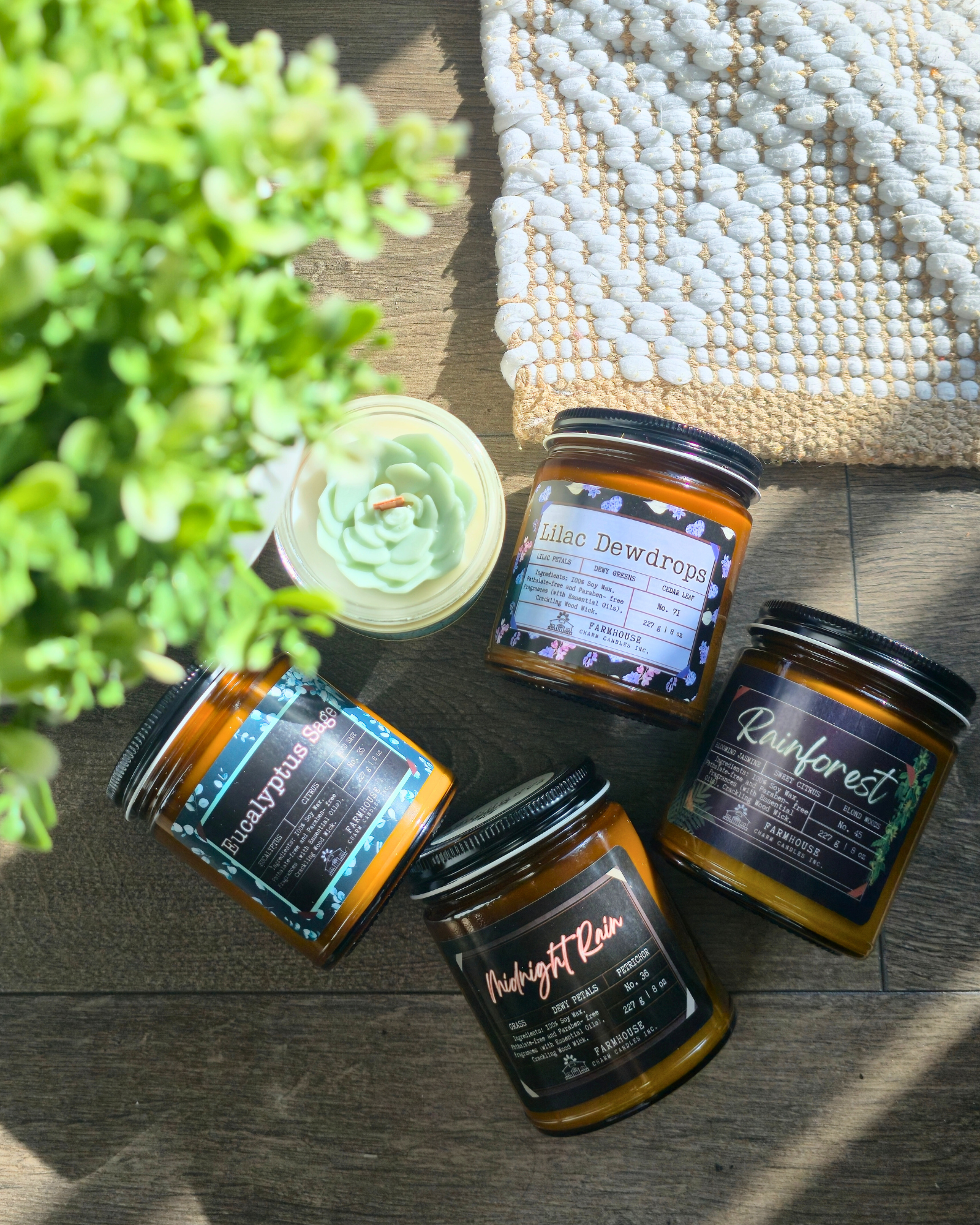 Five spring scented candles with labels on a wooden surface next to a plant and textured fabric. Farmhouse Charm Candles Spring Collection