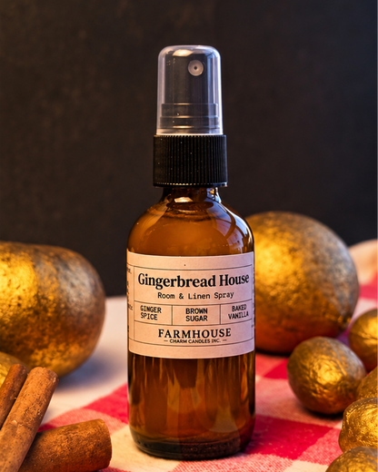 An amber glass room and linen spray bottle with a black spray top and a cream label reading 'Gingerbread House' by Farmhouse Charm Candles, styled with cinnamon sticks and gold apples on a red-and-white check cloth against a dark background.