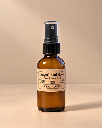 An amber glass spray bottle with a black pump dispenser and a cream-colored label reading 'Gingerbread House Face & Lichen Spray' by Farmhouse Charm Candles set against a warm beige background.